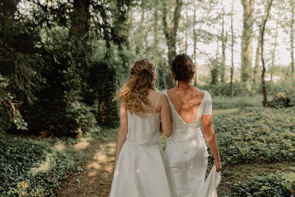 Deux mariées de dos en robe blanche avec des coiffures élégantes lors d'un shooting photo en forêt